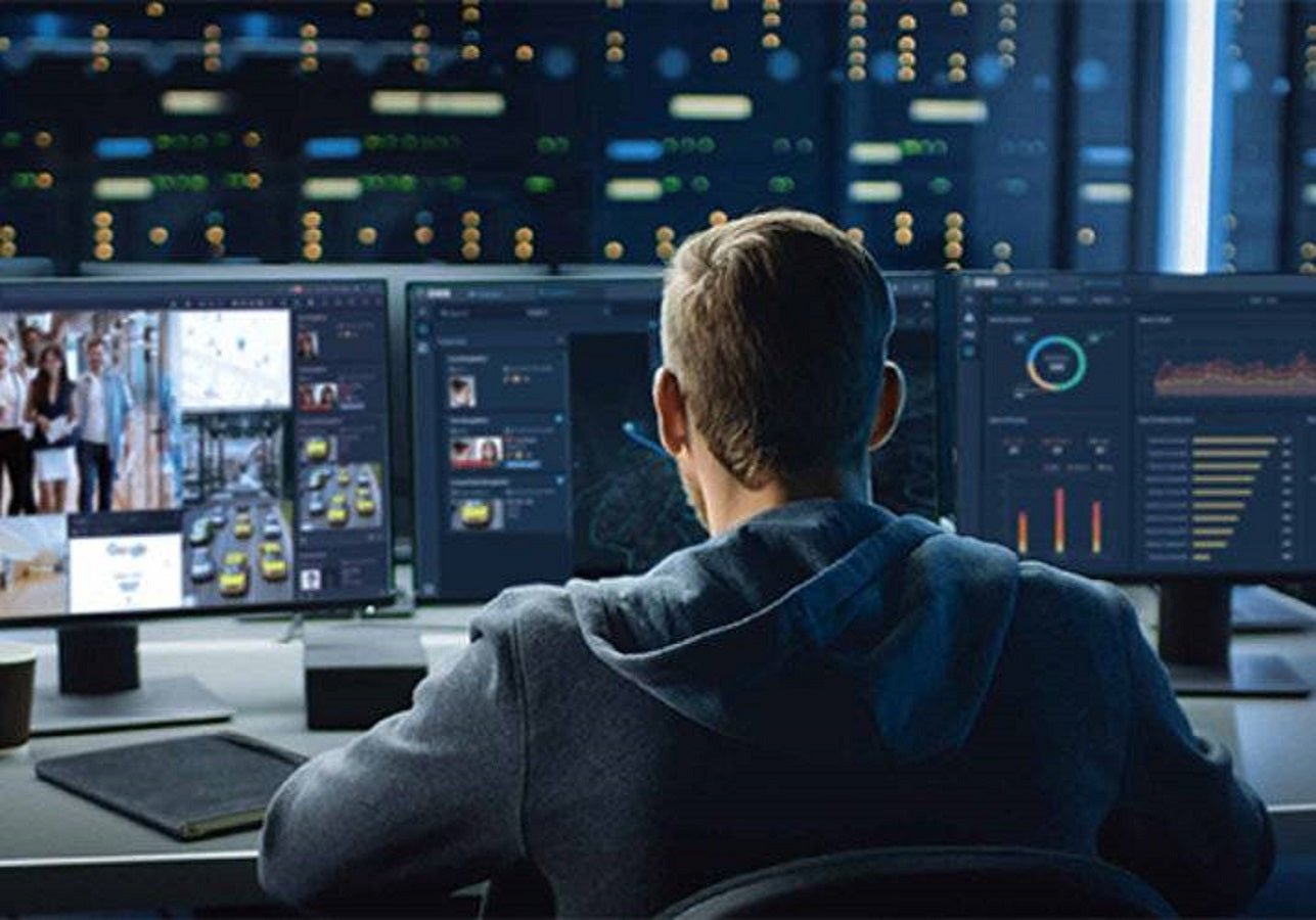 Operador monitoreando sistemas de seguridad, cámaras y datos en sala de control de redes.